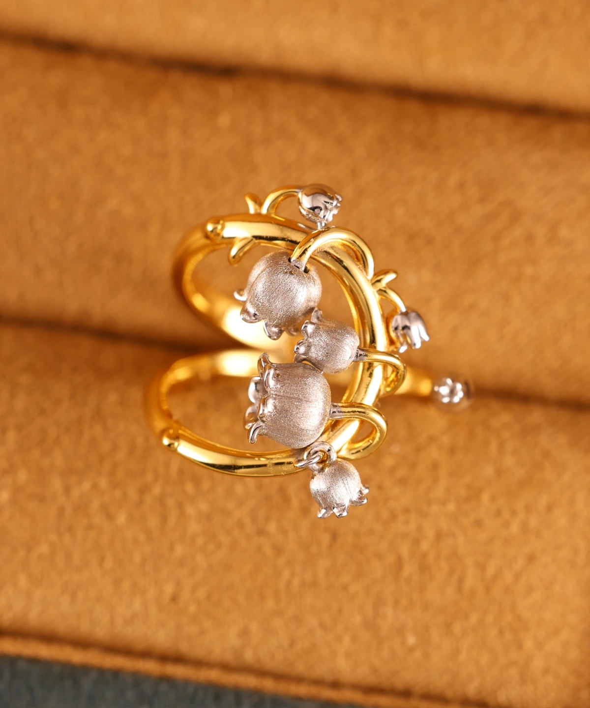 The image shows a beautifully crafted ring named Ethereal Bells | Sterling Silver Lily Of The Valley Ring. The ring features a design inspired by the lily of the valley flower, with delicate, bell-shaped flowers made of sterling silver. These flowers are attached to a gold-plated band that has an intricate, vine-like structure. The combination of the silver flowers and the gold-plated band creates a striking contrast, highlighting the detailed craftsmanship and elegance of the piece. The ring is set against a soft, beige background, which enhances the visual appeal of the jewelry. This Ethereal Bells | Sterling Silver Lily Of The Valley Ring is a stunning example of artistic design and fine materials, making it a desirable addition to any jewelry collection.