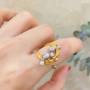 The image features a close-up of a hand wearing the Ethereal Bells | Sterling Silver Lily Of The Valley Ring. The ring is gold-plated with intricate vine-like loops that wrap gracefully around the finger. Adorning the ring are delicate sterling silver elements shaped like lily of the valley flowers, which are small and bell-shaped. The background is softly blurred, ensuring that the focus remains on the ring's detailed craftsmanship and elegant design. This Ethereal Bells | Sterling Silver Lily Of The Valley Ring stands out due to its unique blend of gold-plated and sterling silver elements, making it a stunning and sophisticated accessory.