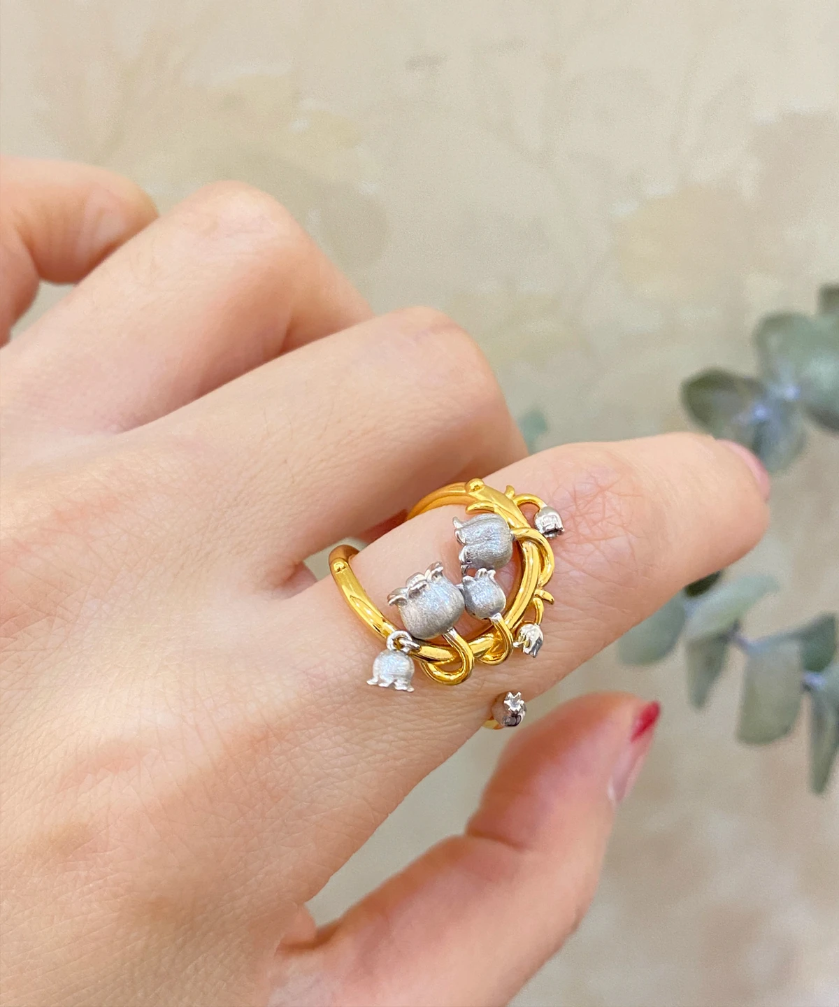 The image features a close-up of a hand wearing the Ethereal Bells | Sterling Silver Lily Of The Valley Ring. The ring is gold-plated with intricate vine-like loops that wrap gracefully around the finger. Adorning the ring are delicate sterling silver elements shaped like lily of the valley flowers, which are small and bell-shaped. The background is softly blurred, ensuring that the focus remains on the ring's detailed craftsmanship and elegant design. This Ethereal Bells | Sterling Silver Lily Of The Valley Ring stands out due to its unique blend of gold-plated and sterling silver elements, making it a stunning and sophisticated accessory.