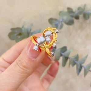 The image features a close-up of a hand wearing the Ethereal Bells | Sterling Silver Lily Of The Valley Ring. The ring is gold-plated with intricate vine-like loops that wrap gracefully around the finger. Adorning the ring are delicate sterling silver elements shaped like lily of the valley flowers, which are small and bell-shaped. The background is softly blurred, ensuring that the focus remains on the ring's detailed craftsmanship and elegant design. This Ethereal Bells | Sterling Silver Lily Of The Valley Ring stands out due to its unique blend of gold-plated and sterling silver elements, making it a stunning and sophisticated accessory.