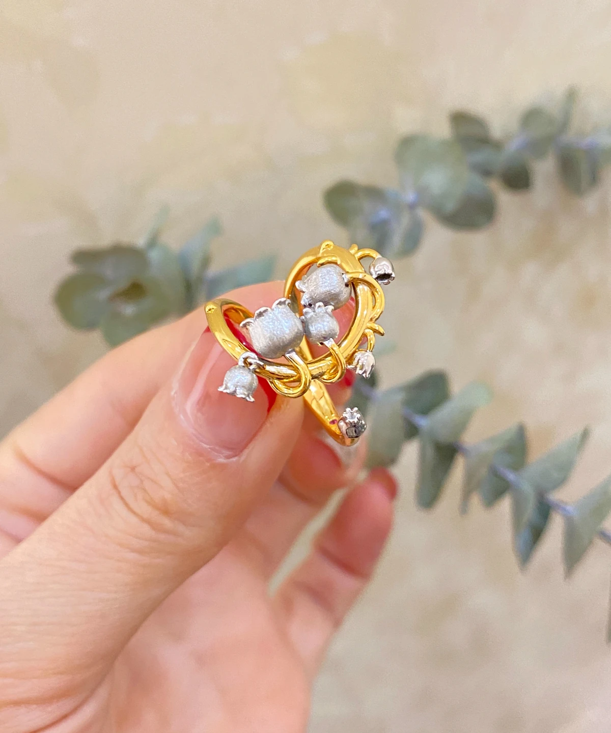 The image features a close-up of a hand wearing the Ethereal Bells | Sterling Silver Lily Of The Valley Ring. The ring is gold-plated with intricate vine-like loops that wrap gracefully around the finger. Adorning the ring are delicate sterling silver elements shaped like lily of the valley flowers, which are small and bell-shaped. The background is softly blurred, ensuring that the focus remains on the ring's detailed craftsmanship and elegant design. This Ethereal Bells | Sterling Silver Lily Of The Valley Ring stands out due to its unique blend of gold-plated and sterling silver elements, making it a stunning and sophisticated accessory.
