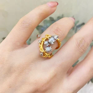 The image features a close-up of a hand wearing the Ethereal Bells | Sterling Silver Lily Of The Valley Ring. The ring is gold-plated with intricate vine-like loops that wrap gracefully around the finger. Adorning the ring are delicate sterling silver elements shaped like lily of the valley flowers, which are small and bell-shaped. The background is softly blurred, ensuring that the focus remains on the ring's detailed craftsmanship and elegant design. This Ethereal Bells | Sterling Silver Lily Of The Valley Ring stands out due to its unique blend of gold-plated and sterling silver elements, making it a stunning and sophisticated accessory.