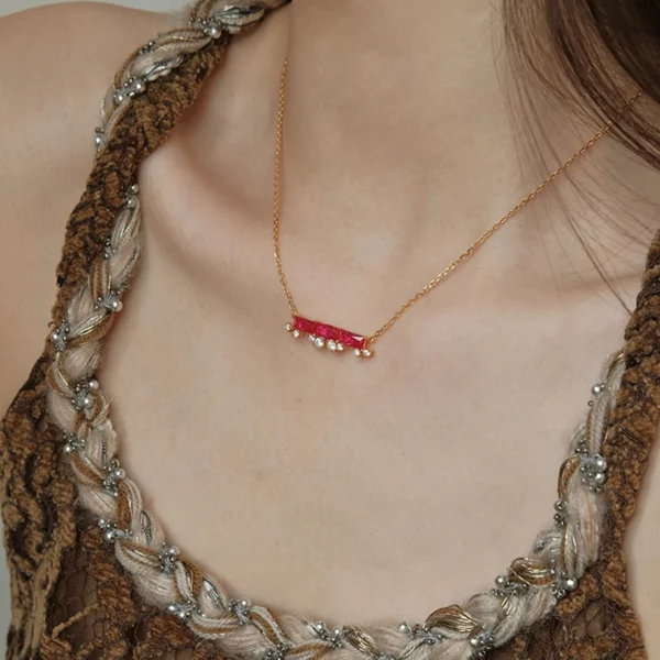 The image shows a close-up view of a model wearing the Rose Realm | Gold Vermeil Three Stone Necklace. This necklace features a gold chain with a horizontal bar pendant that has three rectangular red stones, each set in a gold setting. Below the bar, there are small white pearls or gems hanging, adding an elegant touch to the design. The model is wearing a brown top with intricate braided straps that have silver bead embellishments. The focus of the image is on the necklace and the upper part of the top, highlighting the detailed craftsmanship and sophisticated design of the jewelry.