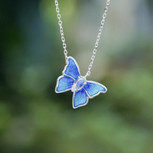 The image shows a close-up view of the Sterling Silver Enamel Blue Butterfly Necklace. The necklace features a delicate butterfly pendant with wings beautifully colored in shades of blue enamel. The wings have a detailed, textured pattern that adds depth and realism to the design. The butterfly's body is made of sterling silver, providing a striking contrast to the vibrant blue wings. The pendant hangs from a fine sterling silver chain with small, evenly spaced links. The background is softly blurred, enhancing the focus on the intricate details and craftsmanship of the butterfly pendant. This Sterling Silver Enamel Blue Butterfly Necklace is a charming and elegant accessory, perfect for adding a touch of nature-inspired beauty to any outfit.