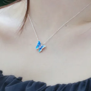 The image shows a close-up view of a model wearing the Sterling Silver Enamel Blue Butterfly Necklace. The necklace features a delicate chain made of sterling silver, with a butterfly-shaped pendant. The butterfly pendant is adorned with blue enamel, creating a vibrant and eye-catching appearance. The model is wearing a dark-colored, ruffled top that contrasts with their light skin tone, highlighting the necklace. This image showcases the Sterling Silver Enamel Blue Butterfly Necklace being worn, providing a clear view of its design and how it looks when worn.