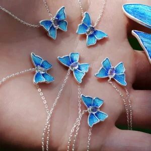 The image shows a close-up view of a hand holding several necklaces, each featuring a butterfly-shaped pendant. The pendants are designed with blue enamel wings, detailed with a textured pattern that adds depth and realism. At the center of each butterfly is a small, clear gemstone. These butterflies are attached to delicate sterling silver chains, making the necklaces visually appealing. The hand is holding six of these necklaces, and there is also a partial view of a blue butterfly-shaped object on the right side of the image. This image effectively showcases the detailed craftsmanship and vibrant blue color of the Sterling Silver Enamel Blue Butterfly Necklace, highlighting its aesthetic appeal.