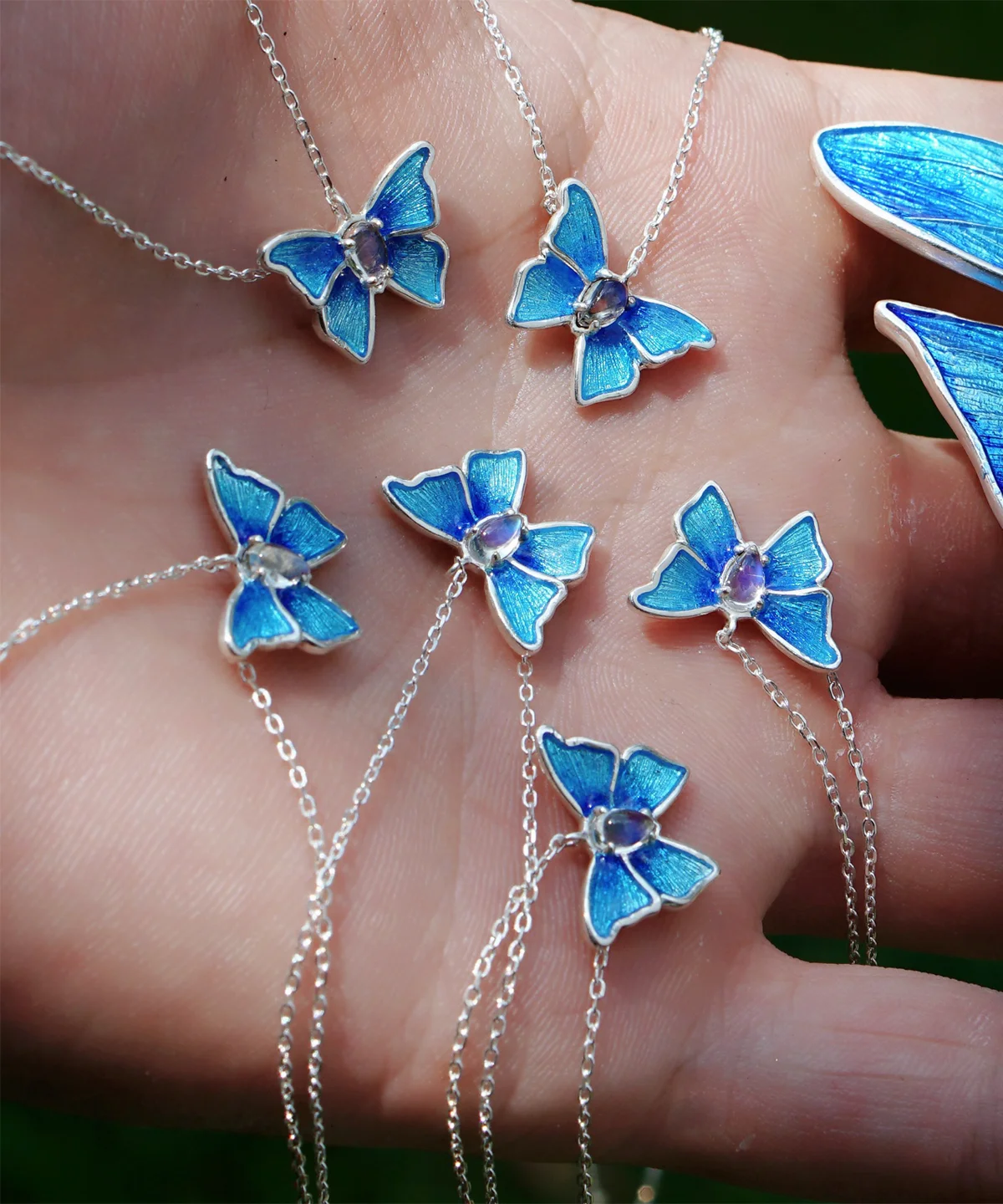 The image shows a close-up view of a hand holding several necklaces, each featuring a butterfly-shaped pendant. The pendants are designed with blue enamel wings, detailed with a textured pattern that adds depth and realism. At the center of each butterfly is a small, clear gemstone. These butterflies are attached to delicate sterling silver chains, making the necklaces visually appealing. The hand is holding six of these necklaces, and there is also a partial view of a blue butterfly-shaped object on the right side of the image. This image effectively showcases the detailed craftsmanship and vibrant blue color of the Sterling Silver Enamel Blue Butterfly Necklace, highlighting its aesthetic appeal.