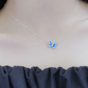 The image shows a close-up view of a model wearing the Sterling Silver Enamel Blue Butterfly Necklace. The necklace features a delicate chain made of sterling silver, with a butterfly-shaped pendant. The butterfly pendant is adorned with blue enamel, creating a vibrant and eye-catching appearance. The model is wearing a dark-colored, ruffled top that contrasts with their light skin tone, highlighting the necklace. This image showcases the Sterling Silver Enamel Blue Butterfly Necklace being worn, providing a clear view of its design and how it looks when worn.