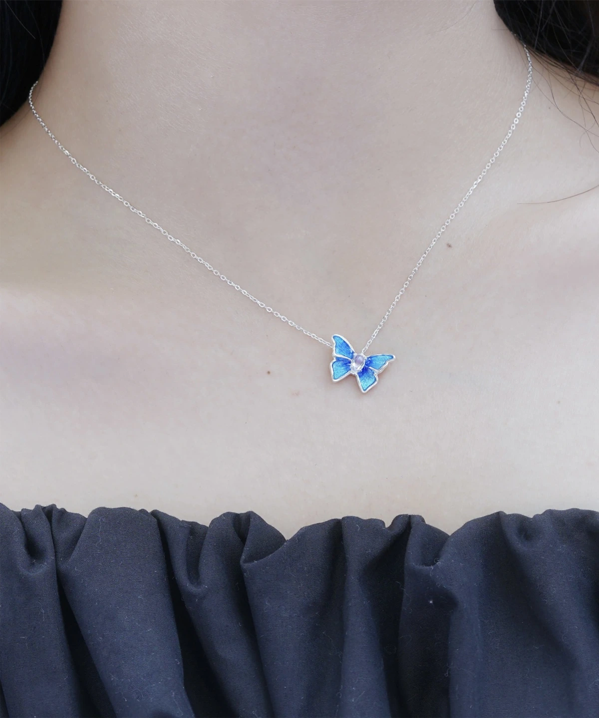 The image shows a close-up view of a model wearing the Sterling Silver Enamel Blue Butterfly Necklace. The necklace features a delicate chain made of sterling silver, with a butterfly-shaped pendant. The butterfly pendant is adorned with blue enamel, creating a vibrant and eye-catching appearance. The model is wearing a dark-colored, ruffled top that contrasts with their light skin tone, highlighting the necklace. This image showcases the Sterling Silver Enamel Blue Butterfly Necklace being worn, providing a clear view of its design and how it looks when worn.