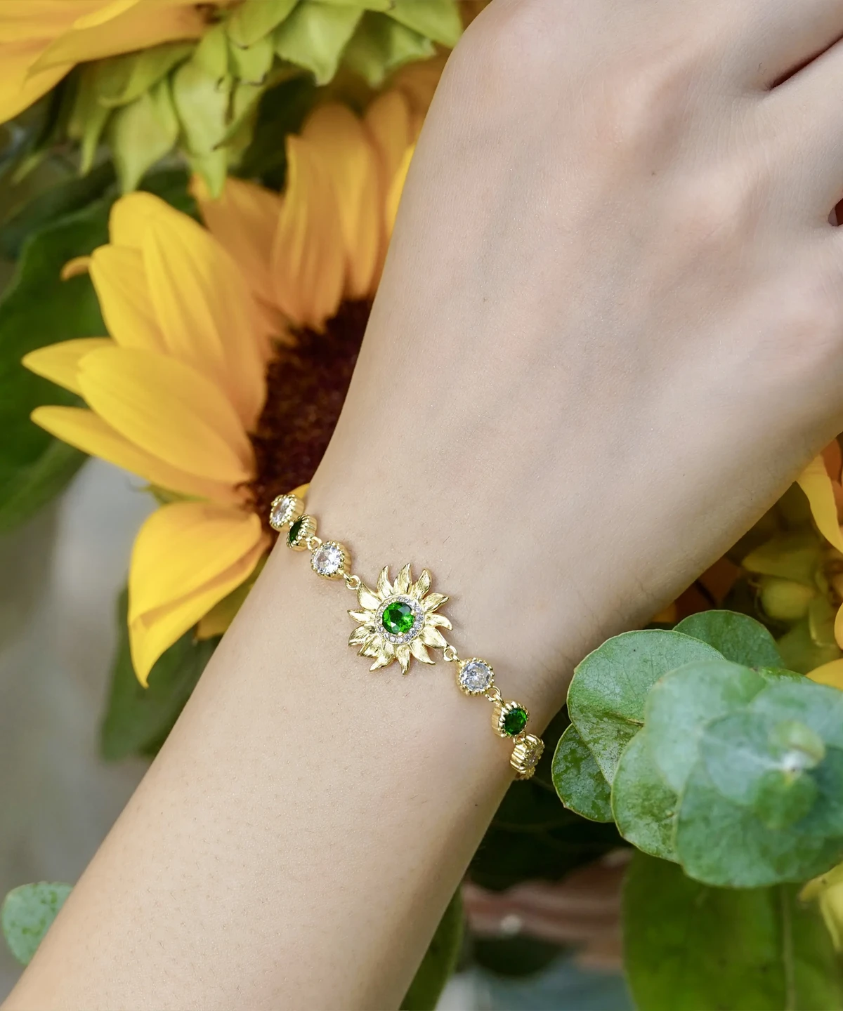 The image shows a close-up of a model wearing the Sterling Silver Sunflower Bracelet. The bracelet features a beautifully crafted sunflower charm with intricate petal detailing and a central green gemstone. The sunflower is attached to a delicate sterling silver chain that wraps elegantly around the wrist. The model's wrist is surrounded by a bouquet of sunflowers and eucalyptus leaves, which complement the sunflower design of the bracelet. The model is wearing a white, textured dress, and the bouquet is wrapped with a blue ribbon. The natural and elegant setting highlights the bracelet's design and how it complements the overall look.