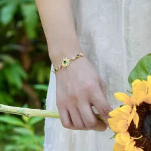 The image shows a close-up of a model wearing the Sterling Silver Sunflower Bracelet. The bracelet features a beautifully crafted sunflower charm with intricate petal detailing and a central green gemstone. The sunflower is attached to a delicate sterling silver chain that wraps elegantly around the wrist. The model's wrist is surrounded by a bouquet of sunflowers and eucalyptus leaves, which complement the sunflower design of the bracelet. The model is wearing a white, textured dress, and the bouquet is wrapped with a blue ribbon. The natural and elegant setting highlights the bracelet's design and how it complements the overall look.