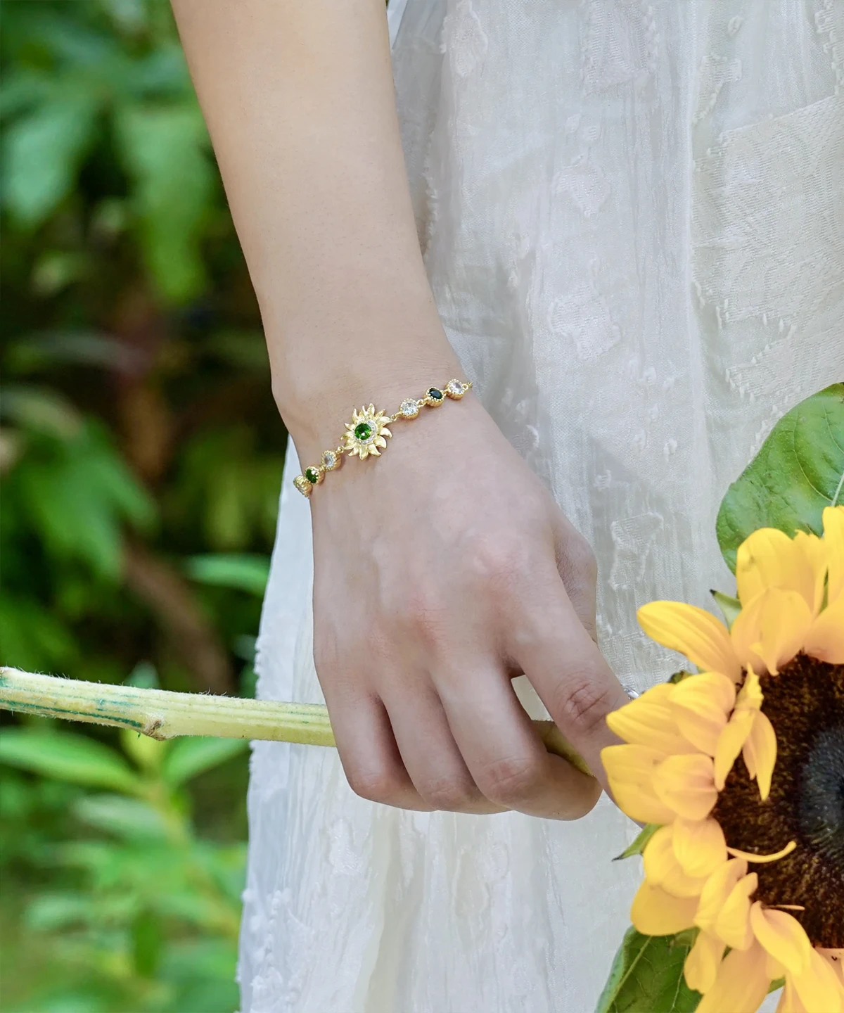 The image shows a close-up of a model wearing the Sterling Silver Sunflower Bracelet. The bracelet features a beautifully crafted sunflower charm with intricate petal detailing and a central green gemstone. The sunflower is attached to a delicate sterling silver chain that wraps elegantly around the wrist. The model's wrist is surrounded by a bouquet of sunflowers and eucalyptus leaves, which complement the sunflower design of the bracelet. The model is wearing a white, textured dress, and the bouquet is wrapped with a blue ribbon. The natural and elegant setting highlights the bracelet's design and how it complements the overall look.