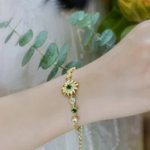 The image shows a close-up of a wrist adorned with the Sterling Silver Sunflower Bracelet. The bracelet features a central sunflower design crafted from sterling silver, with a green gemstone at the center of the sunflower. The bracelet also includes alternating green and clear gemstones set in gold-colored settings along the chain. The model's wrist is positioned gracefully, and the background includes green eucalyptus leaves and a hint of a yellow flower, adding a natural and elegant touch to the overall composition. The intricate design and sparkling details of the bracelet are highlighted, making it stand out as a beautiful and stylish accessory.