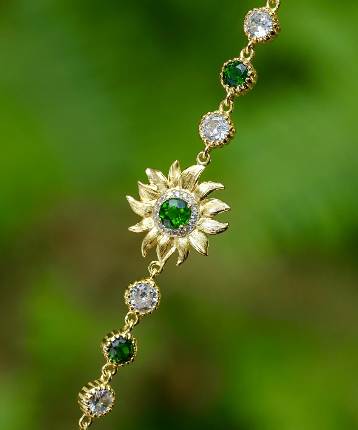 The image shows a close-up view of a beautiful Sterling Silver Sunflower Bracelet. The bracelet features a central sunflower motif, with a finely detailed sunflower crafted from sterling silver. The sunflower has intricate petals radiating outward and a textured center, capturing the essence of the flower. The bracelet chain is made up of small, round links that connect to the sunflower. The background is softly blurred, making the bracelet stand out and highlighting its delicate craftsmanship and elegant design. This Sterling Silver Sunflower Bracelet is a charming accessory that adds a touch of nature-inspired beauty to any outfit.