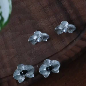 The Butterfly Orchid Earrings—delicate 925 sterling silver floral studs—blend nature-inspired beauty with fine craftsmanship. Each earring features a sculpted orchid with lifelike petals, accented by a black onyx or white pearl at its center for contrast and sophistication. Designed for floral jewelry lovers, these earrings offer an elegant touch to both casual and formal styles. The image presents them on a wooden surface, showcasing their intricate details and polished texture—an exquisite choice for those seeking nature-inspired luxury accessories.
