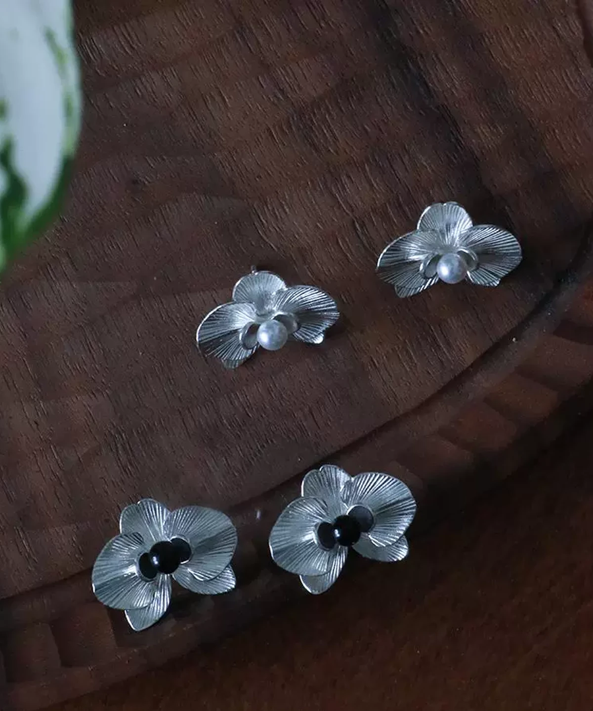 The Butterfly Orchid Earrings—delicate 925 sterling silver floral studs—blend nature-inspired beauty with fine craftsmanship. Each earring features a sculpted orchid with lifelike petals, accented by a black onyx or white pearl at its center for contrast and sophistication. Designed for floral jewelry lovers, these earrings offer an elegant touch to both casual and formal styles. The image presents them on a wooden surface, showcasing their intricate details and polished texture—an exquisite choice for those seeking nature-inspired luxury accessories.