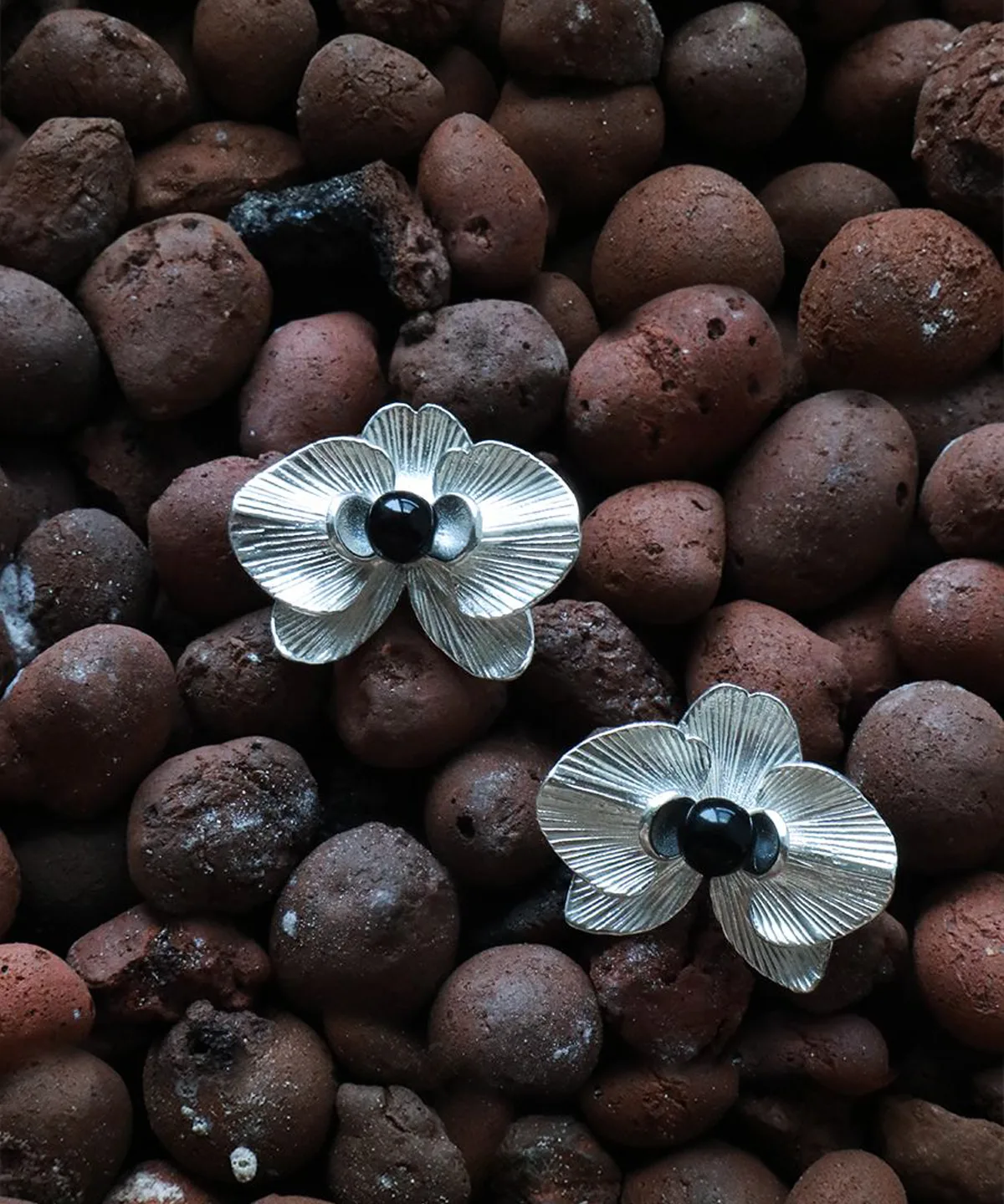 The Butterfly Orchid Earrings—elegant 925 sterling silver floral studs—are inspired by nature’s delicate beauty. Each earring features intricately sculpted orchid petals, with a black onyx centerpiece for contrast and sophistication. Designed for lovers of floral jewelry, these earrings blend fine craftsmanship with timeless elegance. The image showcases them against a textured stone backdrop, emphasizing their refined details and organic inspiration—an ideal accessory for those seeking nature-inspired luxury with a modern touch.