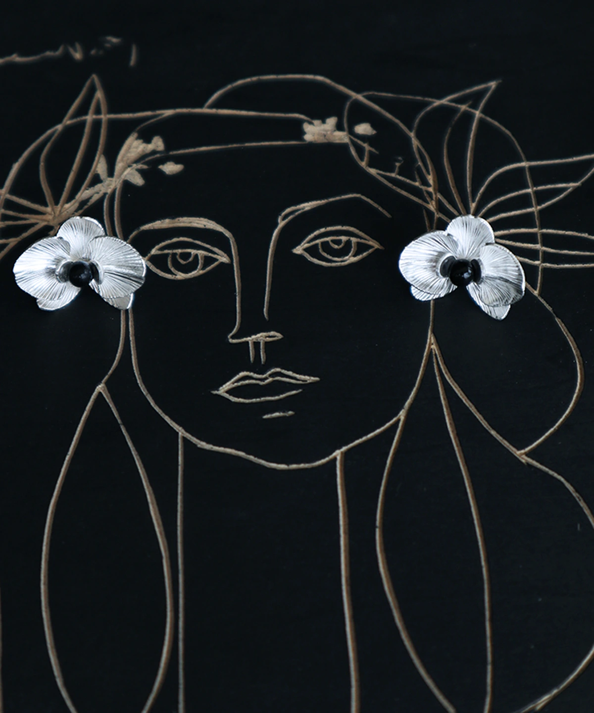 Butterfly Orchid Earrings | 925 Sterling Silver Floral Studs — Displayed on an artistic black canvas with a delicate line-drawn female portrait, these handcrafted earrings resemble blooming orchid petals with intricate textures and a bold black center. The floral studs are placed at the eyes of the portrait, creating a striking visual metaphor. Designed for modern women who appreciate artistic, nature-inspired jewelry with a statement touch. Ideal for gifting or everyday elegance.