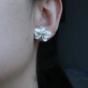 The Butterfly Orchid Earrings—exquisite 925 sterling silver floral studs—feature delicately sculpted orchid petals, with a white pearl at the center, radiating elegance and sophistication. The image captures them worn gracefully, with soft lighting accentuating their polished texture. Set against a neutral backdrop, the earrings’ intricate details and luminous finish stand out. Designed for floral jewelry enthusiasts, these sterling silver studs blend nature-inspired beauty with timeless craftsmanship—perfect for both everyday wear and formal occasions.