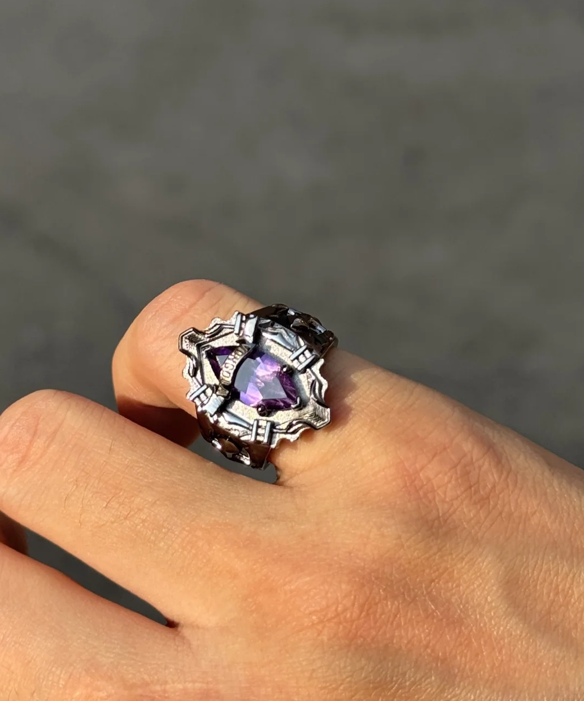 The image features a close-up view of a hand elegantly wearing the Katekyo Hitman Reborn! Vongola Ring. This exquisite piece of Handmade Anime Jewelry is crafted in sterling silver, showcasing a bold and intricate design inspired by Kyoya Hibari’s Vongola Cloud Ring. The ring’s band is detailed with sharp, angular patterns, reflecting the character’s fierce personality and adding a unique, gothic aesthetic. At the center of the ring lies a natural purple amethyst gemstone, radiating a vibrant hue that symbolizes the Cloud Flame’s strength and elegance. The polished silver surface complements the gemstone, emphasizing the artistry of this Katekyo Hitman Reborn Ring. The hand is positioned against a subtle backdrop, highlighting the ring’s craftsmanship and making it a striking accessory for fans and collectors of Vongola Rings