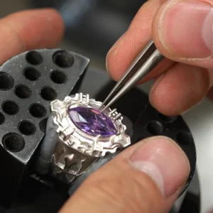 The image captures the precise craftsmanship involved in creating Kyoya Hibari’s Vongola Cloud Ring. This Handmade Anime Jewelry is made of sterling silver and is inspired by the anime Katekyo Hitman Reborn! The photo displays a close-up of the ring, securely held in a vice-like tool on a craftsman’s workbench. The center of the ring features a natural purple amethyst gemstone, which is being carefully adjusted or placed into its setting using a pair of tweezers. The gemstone radiates a vibrant hue, complementing the intricate silver design that embodies the Cloud Flame aesthetic. This unique Katekyo Hitman Reborn Ring showcases the skill and attention to detail required to create iconic Vongola Rings.