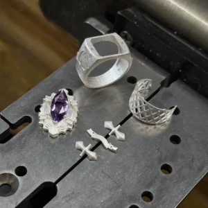 The image captures several components of jewelry in different stages of crafting, highlighting the intricate process behind the creation of Kyoya Hibari’s Vongola Cloud Ring. This piece of Handmade Anime Jewelry is carefully crafted using sterling silver, showcasing attention to detail. Visible in the image are metal elements including a ring with a square design, a piece holding a purple gemstone resembling a natural amethyst, two small cross-shaped components, and a curved lattice-like piece. These parts are arranged on a metal surface, which appears to be part of a workbench or machinery used for jewelry making. This behind-the-scenes look into the production process offers insight into the artistry and craftsmanship involved in creating such unique Katekyo Hitman Reborn Rings and iconic Vongola Rings.