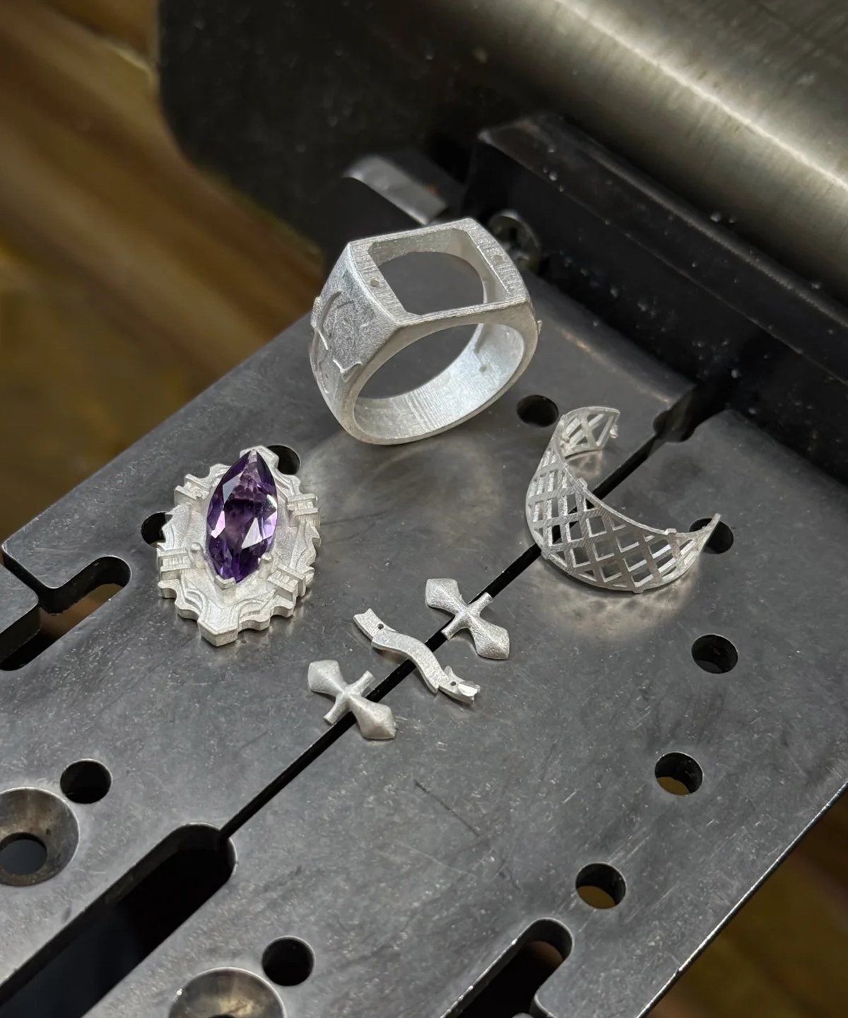 The image captures several components of jewelry in different stages of crafting, highlighting the intricate process behind the creation of Kyoya Hibari’s Vongola Cloud Ring. This piece of Handmade Anime Jewelry is carefully crafted using sterling silver, showcasing attention to detail. Visible in the image are metal elements including a ring with a square design, a piece holding a purple gemstone resembling a natural amethyst, two small cross-shaped components, and a curved lattice-like piece. These parts are arranged on a metal surface, which appears to be part of a workbench or machinery used for jewelry making. This behind-the-scenes look into the production process offers insight into the artistry and craftsmanship involved in creating such unique Katekyo Hitman Reborn Rings and iconic Vongola Rings.