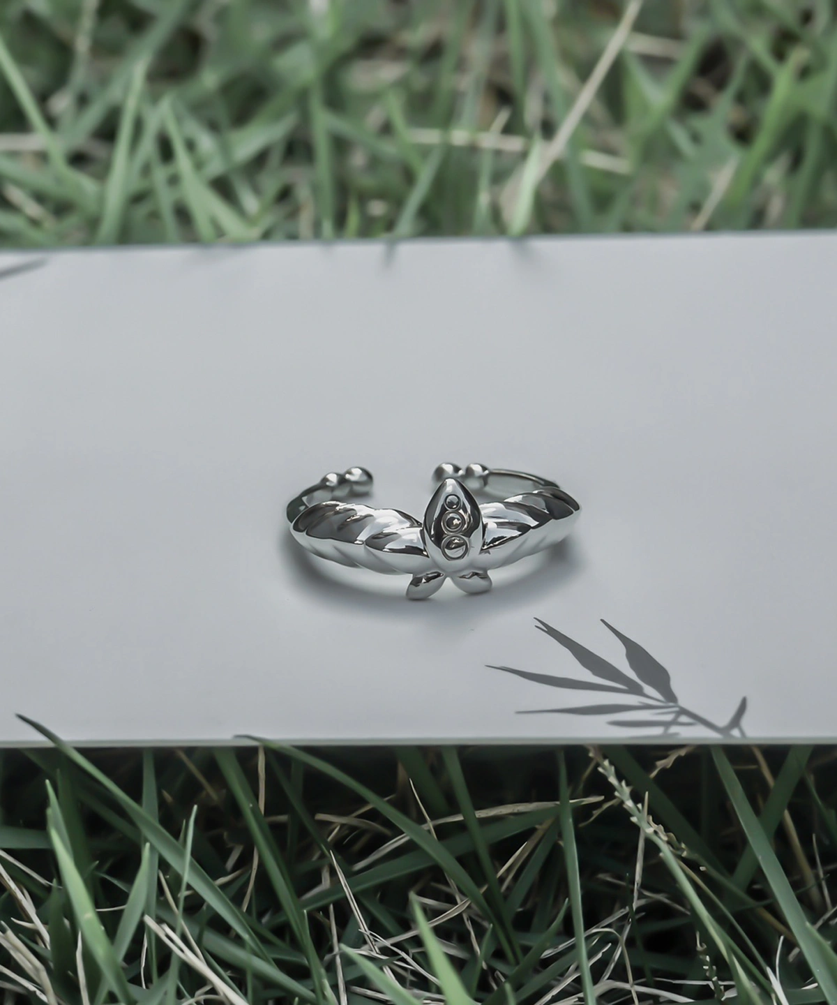 The Mirror Lotus Ring from Lazesoul Handmade Anime Jewelry is a 999 pure silver adjustable ring, inspired by Frieren: Beyond Journey’s End. The image showcases the ring placed on a reflective surface, surrounded by green grass, creating a serene and nature-inspired setting. The intricate lotus motif and mirror-like finish highlight its craftsmanship, making it an ideal choice for anime collectors and jewelry enthusiasts. The open-band design ensures versatile sizing, while the high-quality silver enhances its luminous appeal. A timeless fantasy-inspired accessory, perfect for daily wear or special occasions.