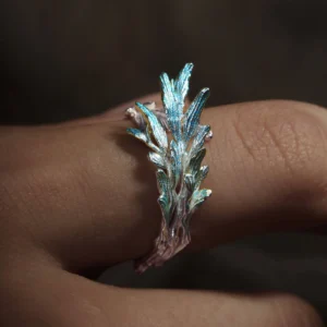 he image showcases a close-up view of the New Leaf | Sterling Silver Open Tree Branch Ring worn on a finger. The ring is designed to resemble a delicate tree branch, with textured detailing along the band that mimics the natural look of bark. Small, polished leaf-shaped motifs extend from the branch, adding elegance and a nature-inspired aesthetic. The sterling silver material has a refined, luminous finish, emphasizing the craftsmanship and organic design. The finger wearing the ring is positioned against a softly blurred background, which highlights the intricate details and sophisticated charm of the New Leaf | Sterling Silver Open Tree Branch Ring.