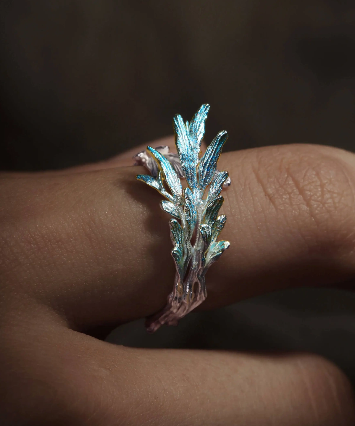 he image showcases a close-up view of the New Leaf | Sterling Silver Open Tree Branch Ring worn on a finger. The ring is designed to resemble a delicate tree branch, with textured detailing along the band that mimics the natural look of bark. Small, polished leaf-shaped motifs extend from the branch, adding elegance and a nature-inspired aesthetic. The sterling silver material has a refined, luminous finish, emphasizing the craftsmanship and organic design. The finger wearing the ring is positioned against a softly blurred background, which highlights the intricate details and sophisticated charm of the New Leaf | Sterling Silver Open Tree Branch Ring.