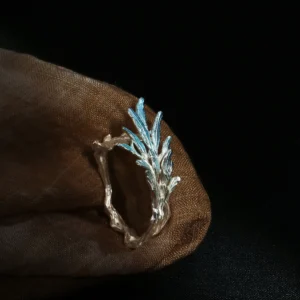 The image showcases an elegant sterling silver tree branch ring named "New Leaf." This ring is crafted to resemble an open tree branch adorned with leaves. The band of the ring is formed by the intertwining branches, giving it a natural and organic appearance. Extending outward from the band are delicate leaves, each featuring a textured surface for added detail. The leaves appear slightly tinted with a blue hue, which complements the silver's gleaming finish. The ring is placed on a warm brown fabric surface, which contrasts beautifully with the dark backdrop. This highlights the ring's intricate design, artistic craftsmanship, and metallic luster. Overall, the "New Leaf" ring embodies a nature-inspired elegance, making it a unique and eye-catching accessory.