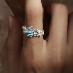 The image features the New Leaf | Sterling Silver Open Tree Branch Ring worn on a finger. The ring is intricately designed to mimic the appearance of a delicate tree branch, with textured detailing on the band to resemble bark. Small, leaf-shaped motifs extend outward from the branch, adding a nature-inspired elegance to the design. The sterling silver material gives the ring a polished, luminous finish. The ring has an open-ended design, offering adjustability for a comfortable fit. The background is softly blurred, emphasizing the fine craftsmanship and unique aesthetic of the ring. This image beautifully showcases the organic and artistic design of the New Leaf | Sterling Silver Open Tree Branch Ring.