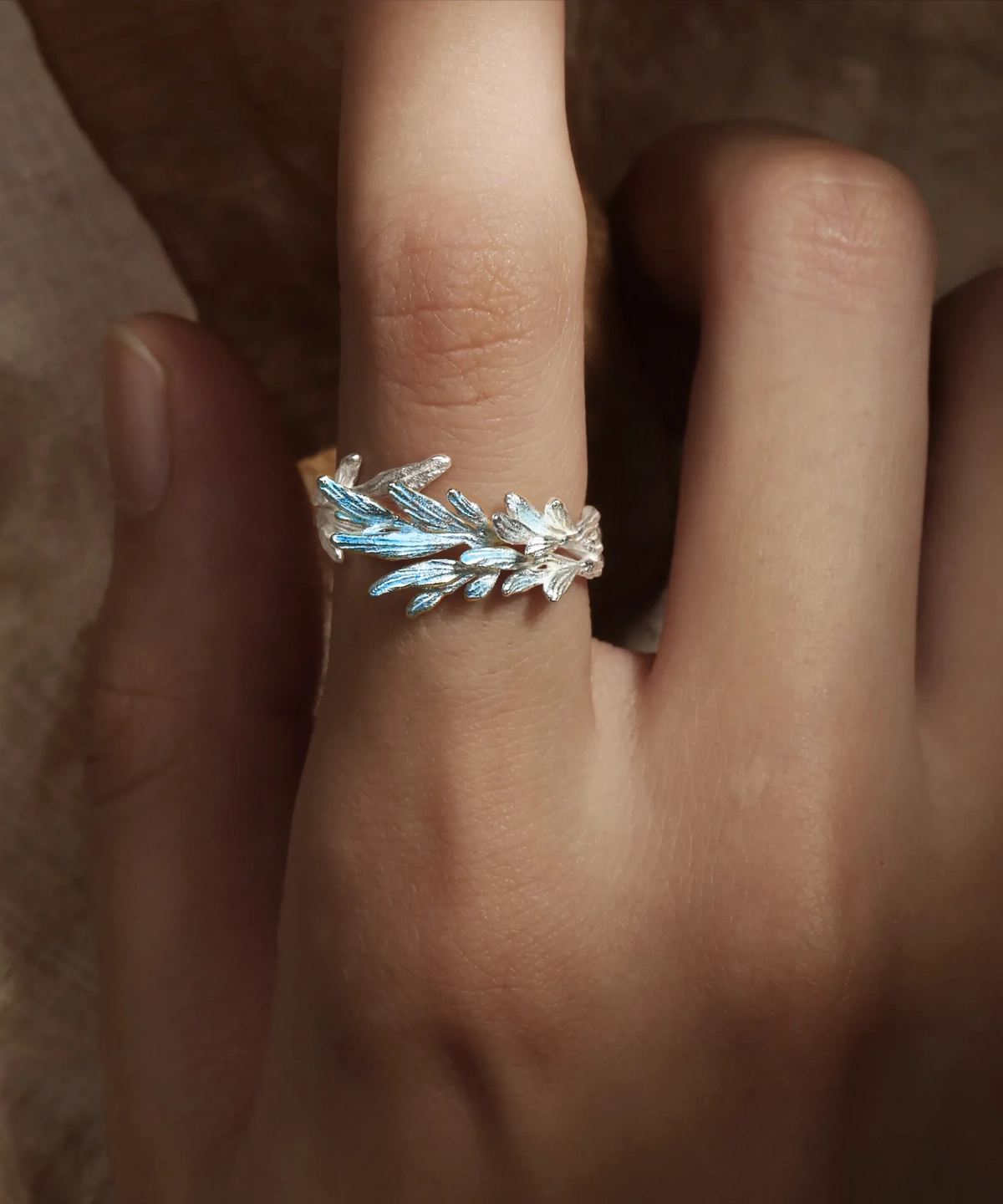 The image features the New Leaf | Sterling Silver Open Tree Branch Ring worn on a finger. The ring is intricately designed to mimic the appearance of a delicate tree branch, with textured detailing on the band to resemble bark. Small, leaf-shaped motifs extend outward from the branch, adding a nature-inspired elegance to the design. The sterling silver material gives the ring a polished, luminous finish. The ring has an open-ended design, offering adjustability for a comfortable fit. The background is softly blurred, emphasizing the fine craftsmanship and unique aesthetic of the ring. This image beautifully showcases the organic and artistic design of the New Leaf | Sterling Silver Open Tree Branch Ring.