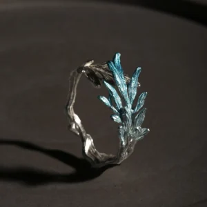 A delicate sterling silver open ring designed to resemble a natural tree branch, featuring a textured, organic band that wraps partially around the finger. One end of the ring is adorned with a cluster of finely detailed leaves that extend upward, finished in a subtle metallic blue-silver hue, creating a striking contrast against the matte silver branch. The overall design is airy and asymmetrical, evoking the fresh growth of new leaves on a branch, and the ring is photographed against a dark, softly lit background to highlight its intricate craftsmanship and natural inspiration.