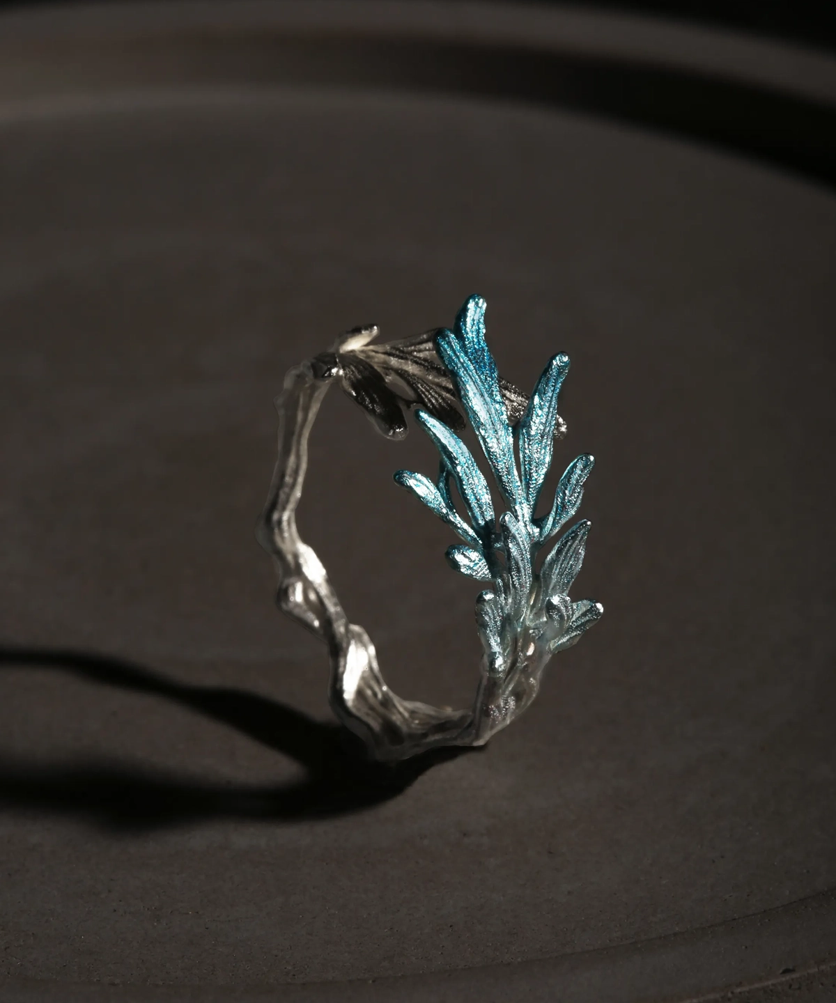 A delicate sterling silver open ring designed to resemble a natural tree branch, featuring a textured, organic band that wraps partially around the finger. One end of the ring is adorned with a cluster of finely detailed leaves that extend upward, finished in a subtle metallic blue-silver hue, creating a striking contrast against the matte silver branch. The overall design is airy and asymmetrical, evoking the fresh growth of new leaves on a branch, and the ring is photographed against a dark, softly lit background to highlight its intricate craftsmanship and natural inspiration.