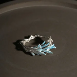 A delicate sterling silver open ring designed to resemble a natural tree branch, featuring a textured, organic band that wraps partially around the finger. One end of the ring is adorned with a cluster of finely detailed leaves that extend upward, finished in a subtle metallic blue-silver hue, creating a striking contrast against the matte silver branch. The overall design is airy and asymmetrical, evoking the fresh growth of new leaves on a branch, and the ring is photographed against a dark, softly lit background to highlight its intricate craftsmanship and natural inspiration.