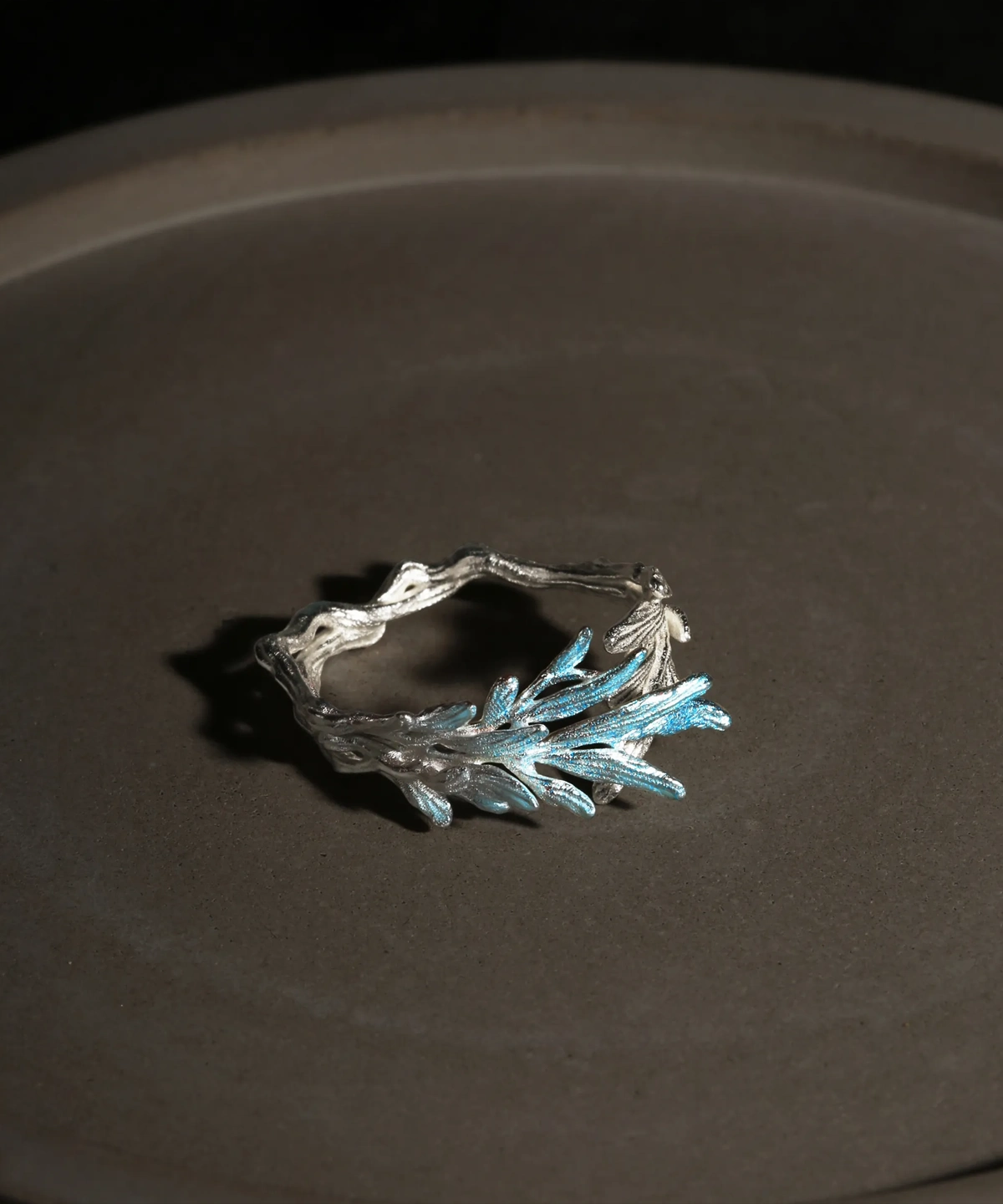 A delicate sterling silver open ring designed to resemble a natural tree branch, featuring a textured, organic band that wraps partially around the finger. One end of the ring is adorned with a cluster of finely detailed leaves that extend upward, finished in a subtle metallic blue-silver hue, creating a striking contrast against the matte silver branch. The overall design is airy and asymmetrical, evoking the fresh growth of new leaves on a branch, and the ring is photographed against a dark, softly lit background to highlight its intricate craftsmanship and natural inspiration.