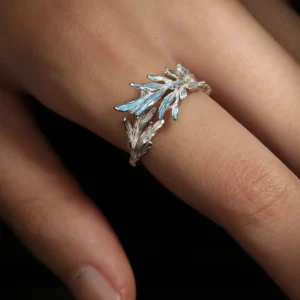 The image features the New Leaf | Sterling Silver Open Tree Branch Ring worn on a finger. The ring is intricately designed to mimic the appearance of a delicate tree branch, with textured detailing on the band to resemble bark. Small, leaf-shaped motifs extend outward from the branch, adding a nature-inspired elegance to the design. The sterling silver material gives the ring a polished, luminous finish. The ring has an open-ended design, offering adjustability for a comfortable fit. The background is softly blurred, emphasizing the fine craftsmanship and unique aesthetic of the ring. This image beautifully showcases the organic and artistic design of the New Leaf | Sterling Silver Open Tree Branch Ring.