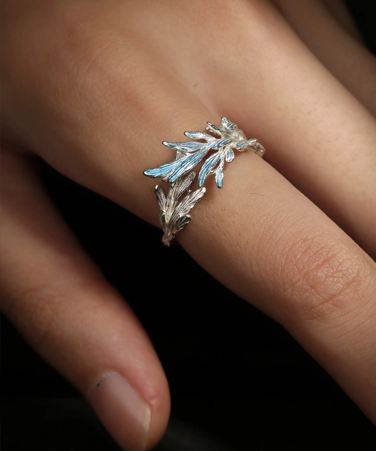 The image features the New Leaf | Sterling Silver Open Tree Branch Ring worn on a finger. The ring is intricately designed to mimic the appearance of a delicate tree branch, with textured detailing on the band to resemble bark. Small, leaf-shaped motifs extend outward from the branch, adding a nature-inspired elegance to the design. The sterling silver material gives the ring a polished, luminous finish. The ring has an open-ended design, offering adjustability for a comfortable fit. The background is softly blurred, emphasizing the fine craftsmanship and unique aesthetic of the ring. This image beautifully showcases the organic and artistic design of the New Leaf | Sterling Silver Open Tree Branch Ring.