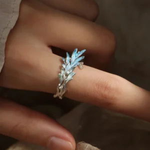 The image features the New Leaf | Sterling Silver Open Tree Branch Ring worn on a finger. The ring is intricately designed to mimic the appearance of a delicate tree branch, with textured detailing on the band to resemble bark. Small, leaf-shaped motifs extend outward from the branch, adding a nature-inspired elegance to the design. The sterling silver material gives the ring a polished, luminous finish. The ring has an open-ended design, offering adjustability for a comfortable fit. The background is softly blurred, emphasizing the fine craftsmanship and unique aesthetic of the ring. This image beautifully showcases the organic and artistic design of the New Leaf | Sterling Silver Open Tree Branch Ring.