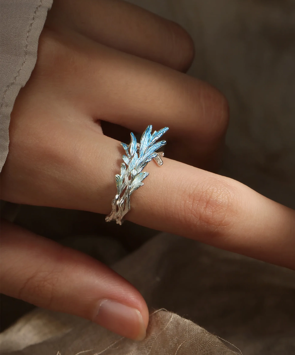The image features the New Leaf | Sterling Silver Open Tree Branch Ring worn on a finger. The ring is intricately designed to mimic the appearance of a delicate tree branch, with textured detailing on the band to resemble bark. Small, leaf-shaped motifs extend outward from the branch, adding a nature-inspired elegance to the design. The sterling silver material gives the ring a polished, luminous finish. The ring has an open-ended design, offering adjustability for a comfortable fit. The background is softly blurred, emphasizing the fine craftsmanship and unique aesthetic of the ring. This image beautifully showcases the organic and artistic design of the New Leaf | Sterling Silver Open Tree Branch Ring.