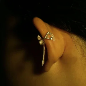 Close-up view of our 925 Sterling Silver Bow Ear Cuff | No Piercing Earrings worn on the ear's backside. This artistic ear cuff features a delicate, bow-inspired design with intricate details reminiscent of a small branch adorned with subtle leaf accents and a dangling motif. The luminous silver finish contrasts beautifully with the dark backdrop, emphasizing its refined craftsmanship and modern elegance—ideal for fashion-forward individuals seeking stylish, non-piercing ear jewelry.