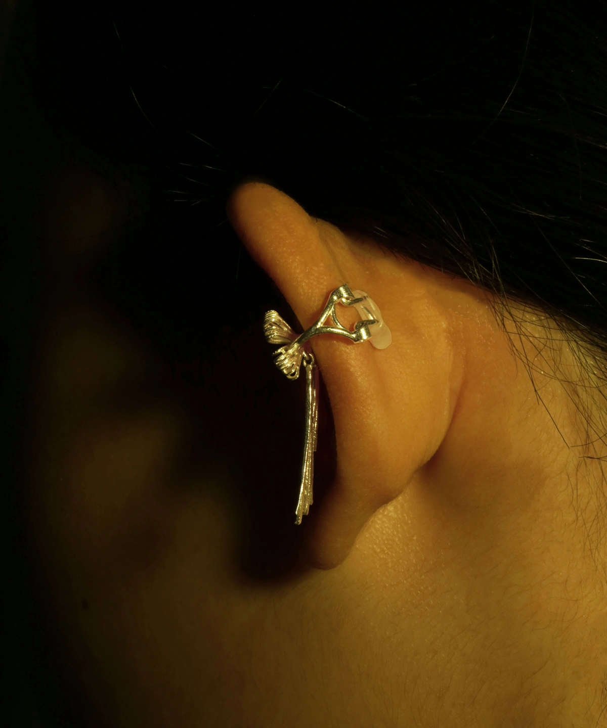 Close-up view of our 925 Sterling Silver Bow Ear Cuff | No Piercing Earrings worn on the ear's backside. This artistic ear cuff features a delicate, bow-inspired design with intricate details reminiscent of a small branch adorned with subtle leaf accents and a dangling motif. The luminous silver finish contrasts beautifully with the dark backdrop, emphasizing its refined craftsmanship and modern elegance—ideal for fashion-forward individuals seeking stylish, non-piercing ear jewelry.