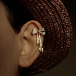 Close-up view of our 925 Sterling Silver Bow Ear Cuff | No Piercing Earrings. This ear cuff features an intricate bow design with delicate cascading strands, elegantly worn on the mid-ear. The image, set against a backdrop partially revealed by a textured woven hat and bathed in warm, dim lighting, emphasizes its refined craftsmanship. Ideal for fashion-forward individuals, this non-piercing accessory adds a touch of modern sophistication and timeless elegance to both everyday and special occasion looks.