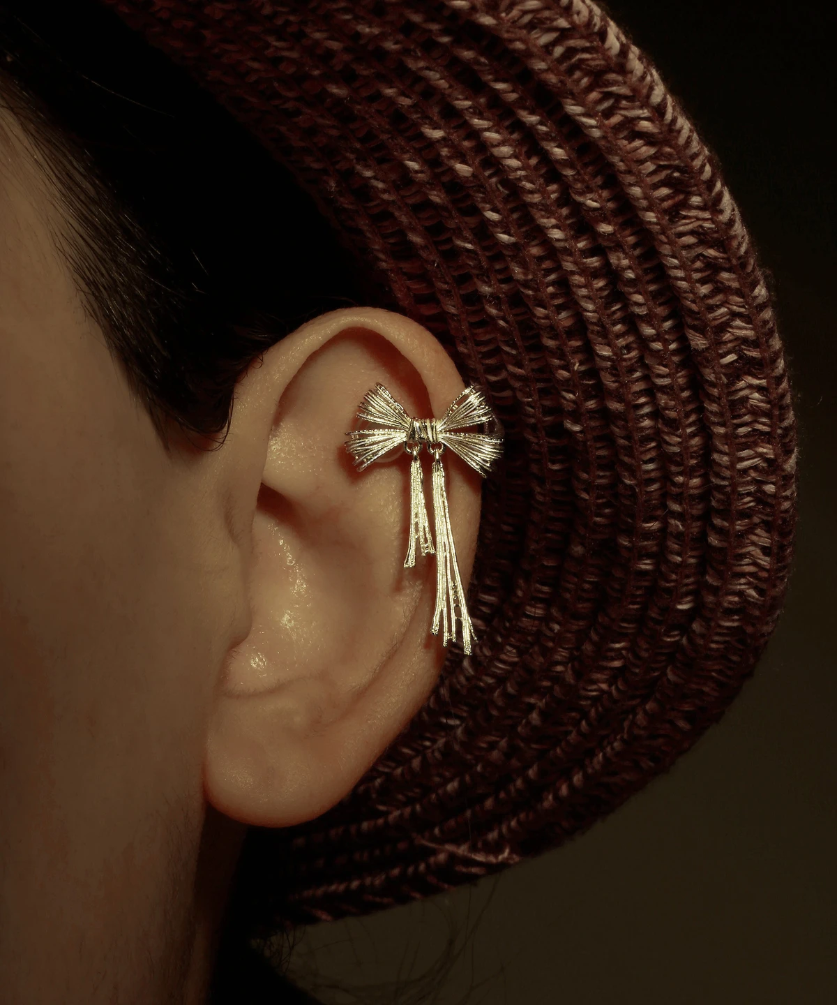 Close-up view of our 925 Sterling Silver Bow Ear Cuff | No Piercing Earrings. This ear cuff features an intricate bow design with delicate cascading strands, elegantly worn on the mid-ear. The image, set against a backdrop partially revealed by a textured woven hat and bathed in warm, dim lighting, emphasizes its refined craftsmanship. Ideal for fashion-forward individuals, this non-piercing accessory adds a touch of modern sophistication and timeless elegance to both everyday and special occasion looks.