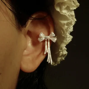 This close-up image of our 925 Sterling Silver Bow Ear Cuff | No Piercing Earrings shows the accessory worn on the middle part of the ear. The ear cuff features an intricately crafted bow design with delicate detailing, exemplifying modern elegance and timeless charm. Set against a soft lace-textured backdrop, the photo highlights its artisanal quality and wearable style. Ideal for fashion-forward individuals seeking statement jewelry without piercings, this ear cuff balances sophisticated design with everyday versatility.