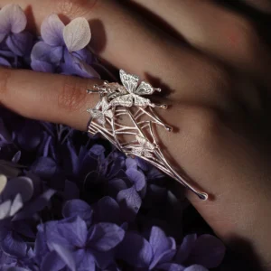 The image showcases our Butterfly Ring in 925 Sterling Silver, an Art Fantasy Pinky Ring featuring intricate butterfly motifs and delicate web detailing. Worn on a model’s pinky against a vibrant backdrop of purple blooms, the ring exudes whimsical artistry and refined craftsmanship. Ideal for both casual and formal looks, this enchanting accessory adds a touch of fantasy and sophistication to any outfit.