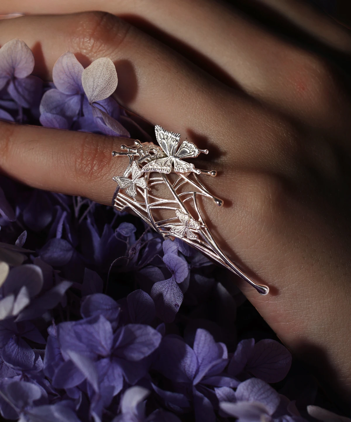 The image showcases our Butterfly Ring in 925 Sterling Silver, an Art Fantasy Pinky Ring featuring intricate butterfly motifs and delicate web detailing. Worn on a model’s pinky against a vibrant backdrop of purple blooms, the ring exudes whimsical artistry and refined craftsmanship. Ideal for both casual and formal looks, this enchanting accessory adds a touch of fantasy and sophistication to any outfit.