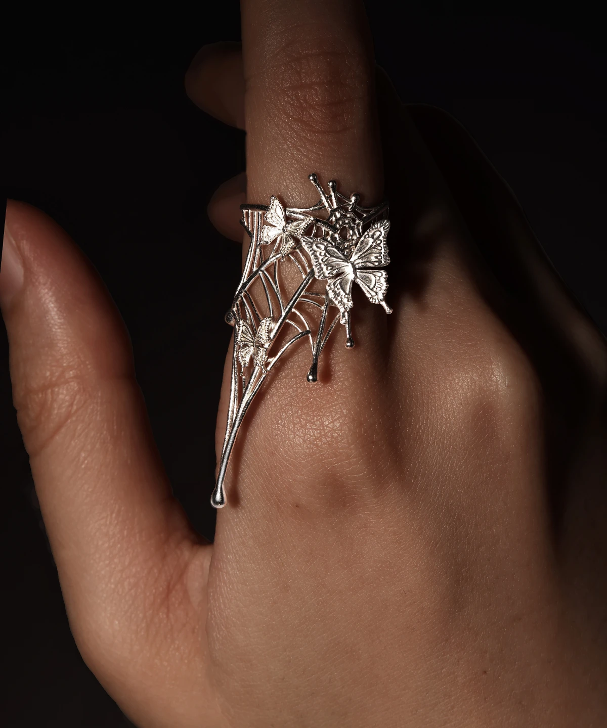 Close-up image of a hand wearing our Butterfly Ring in 925 Sterling Silver | Art Fantasy Pinky Ring. Featuring an intricate, web-inspired design adorned with delicate butterfly motifs, this ring highlights exceptional craftsmanship. The model's hand is set against a dark, textured backdrop, emphasizing the ring’s artistic detail and bold allure. Perfect for fashion-forward trendsetters seeking whimsical, statement jewelry, this unique pinky ring seamlessly blends modern elegance with fantasy-inspired design.