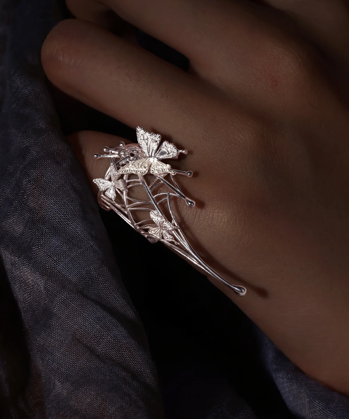 Butterfly Ring in 925 Sterling Silver | Art Fantasy Pinky Ring features an intricately designed silver band adorned with delicate butterfly motifs and a subtle web-inspired pattern. Captured in a close-up on a model’s hand against a dark, textured backdrop, the ring’s shimmering details stand out beautifully. This whimsical yet elegant piece exudes refined craftsmanship, making it a perfect statement accessory for both casual and formal wear, ideal for fashion-forward trendsetters.